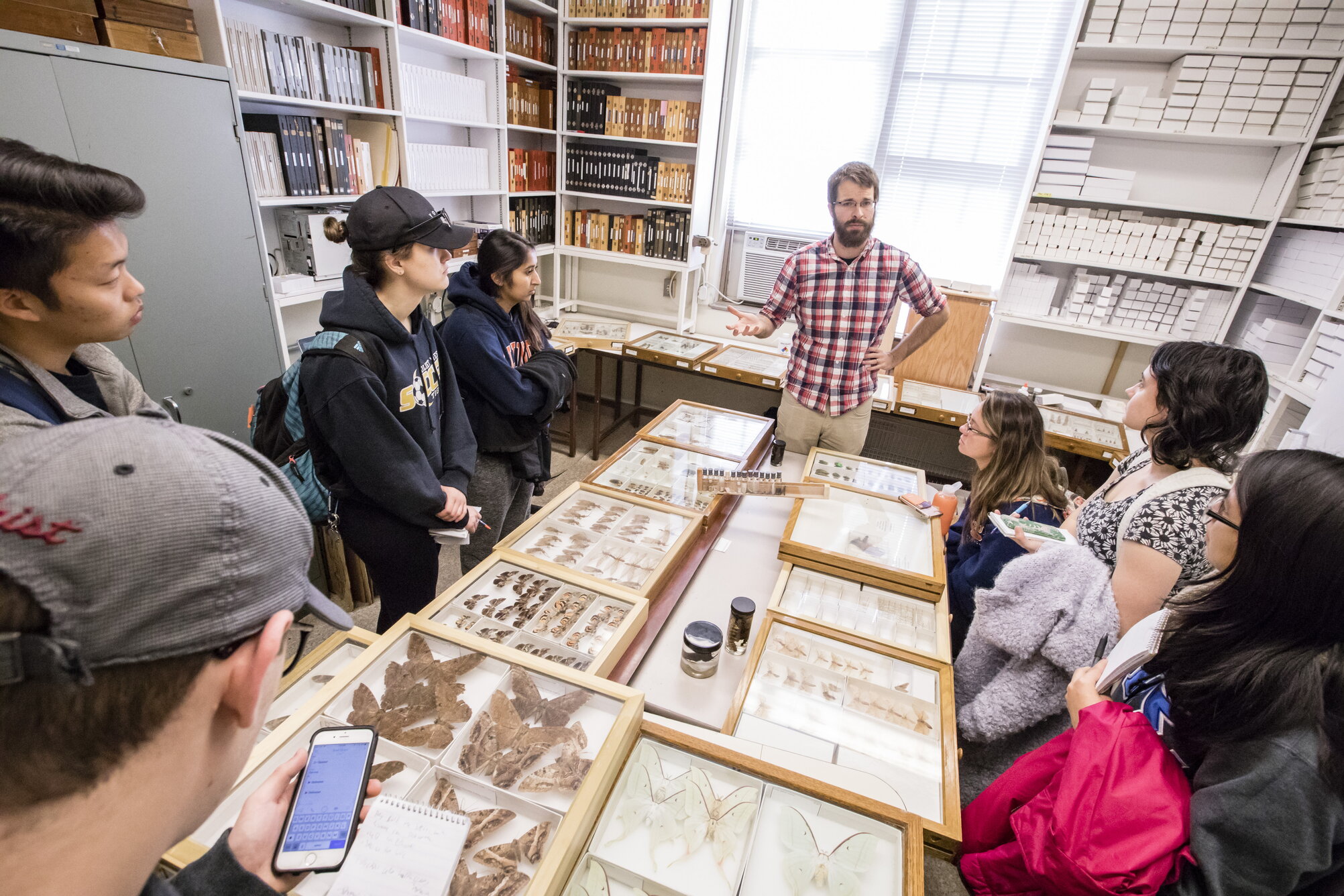 A person giving a lecture to students gathered around insect collections.