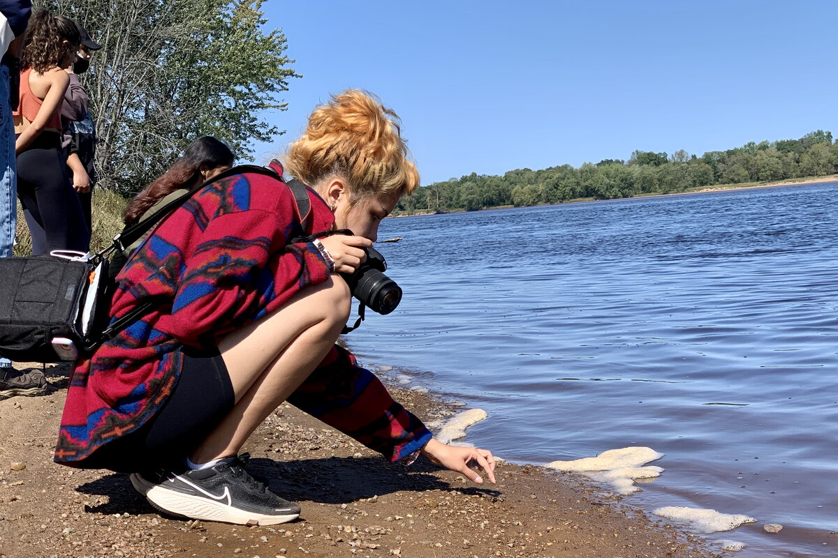 A student taking a picture of a rock next to a lake.